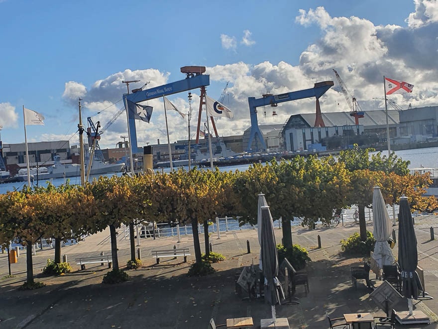 Blick auf der Kieler Förde, vorne Bäume mit Herbstlaub, hinten der Werft, blauer Himmel dramatische weiße Wolken
