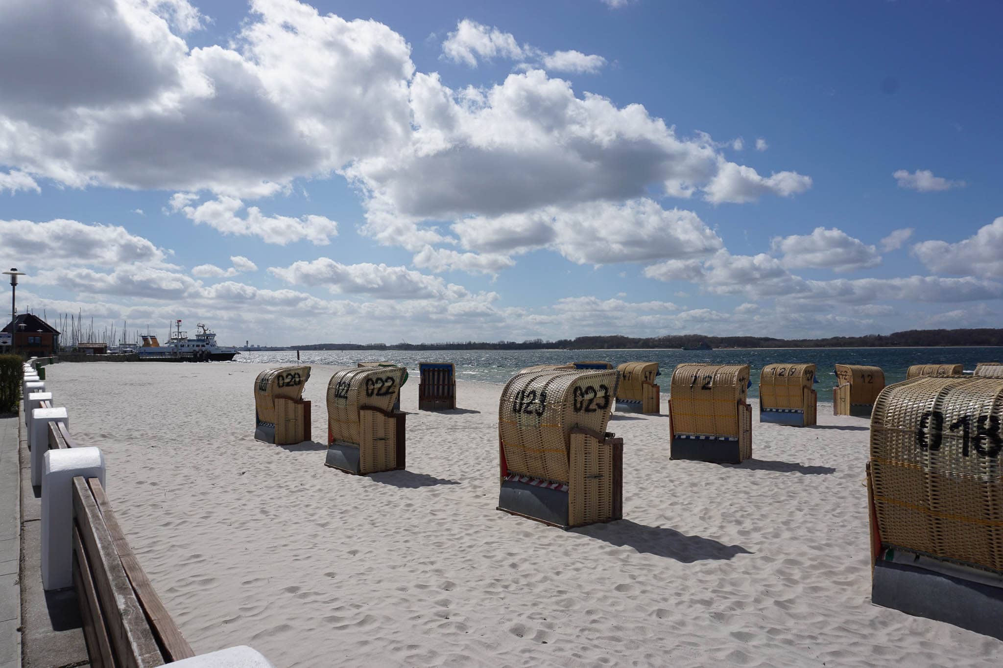 Im Vordergrund Strand und Strandkörbe, im Hintergrund der Kieler Förde und blauer Himmel mit weiße Wölkchen